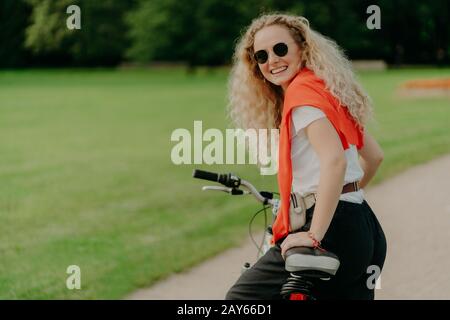 Heureuse jeune femme avec des cheveux frisés, regarde de l'arrière, s'arrête pendant la bicyclette de conduite, attend pour un ami qui est derrière, porte des lunettes de soleil, chandail sur l'épaule Banque D'Images