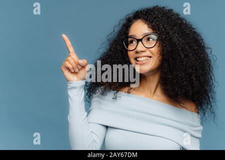 Une femme sourit et sourit avec une peau sombre et heureuse, avec des points de coiffure Afro, index finger vers le haut, montre quelque chose sur l'espace vide, a une expression heureuse, porte Banque D'Images