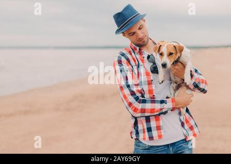 Le tir en plein air d'un bel homme porte un chapeau et une chemise à damier, porte votre chien préféré dans les mains, se promener sur la côte pendant la journée d'été, ont r amical Banque D'Images