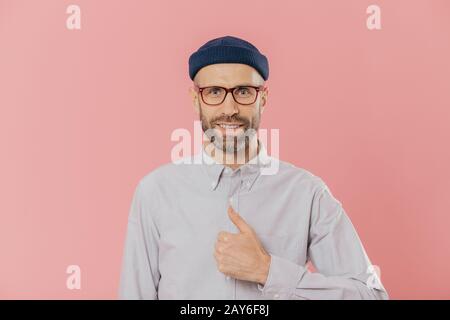 Satisfait l'homme a beau look attrayant, soulève le pouce, d'accord avec quelque chose, sourit joyeusement, porte un chapeau et chemise, plus isolé sur fond rose. Banque D'Images