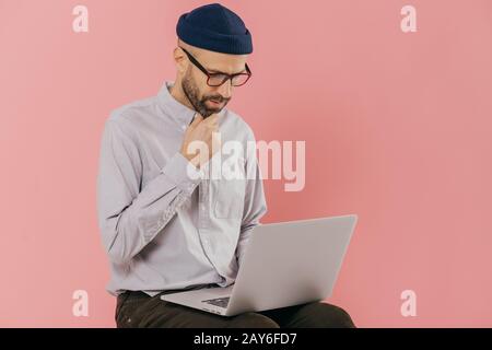 Photo de l'homme non rasé concentré est titulaire d'chin, regarde attentivement à l'écran d'ordinateur portable, bénéficie d communication en ligne, porte des lunettes et optique Banque D'Images