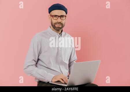 Homme graphic designer réfléchit à des idées créatives, se lit l'actualité des site internet, utilise un ordinateur portable moderne, est assis contre fond rose. Business Banque D'Images