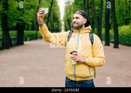 Portrait isolé de type hipster portant des vêtements tendance reposant dans le parc seul en admirant le bon temps et l'air frais. Homme barbu faisant selfie dans p Banque D'Images