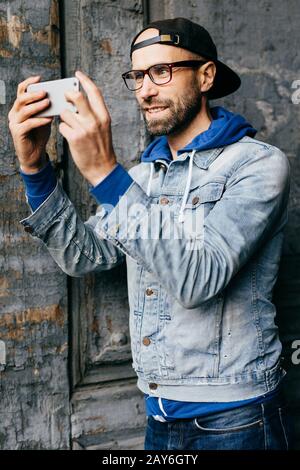 Portrait d'un type élégant avec barbe portant des vêtements tendance tenant le téléphone mobile, rendant le selfie heureux et souriant avec joie dans l'appareil photo. Détendu m Banque D'Images