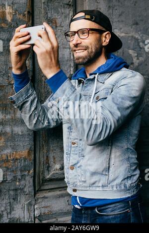 Un homme excité avec barbe et moustache portant une chemise denim tendance avec un crochet et un Jean regardant avec plaisir dans l'appareil photo de son smartphone qui fait du selfie. Fond Banque D'Images