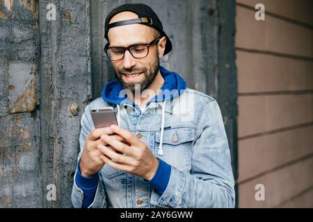 Portrait horizontal de l'homme élégant barbu en denim anorak portant de grandes lunettes tenant smartphone à l'aide d'Internet gratuit, en vérifiant l'e-mail, wa Banque D'Images