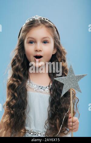 Petite fille princesse féminine porte couronne et robe, tient baguette magique, a de longs cheveux cally foncé pose sur fond bleu. Beau preaprre de gamin Banque D'Images