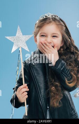 Un petit enfant positif et surémotif couvre la bouche avec une paume, porte une veste et une couronne en cuir noir tendance, tient la baguette magique dans la main, isolée au-dessus Banque D'Images