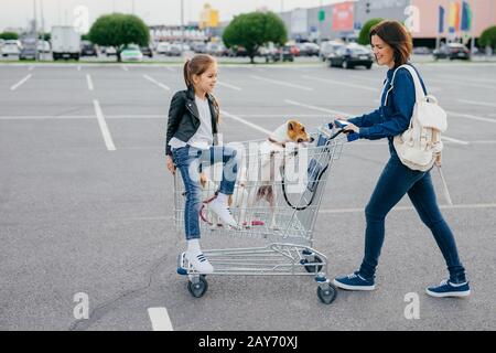 La photo horizontale d'une femme heureuse porte un sac à dos, vêtu de vêtements en denim, tient une voiturette, sa petite fille et deux chiens posent dans un chariot, retour à la maison A Banque D'Images