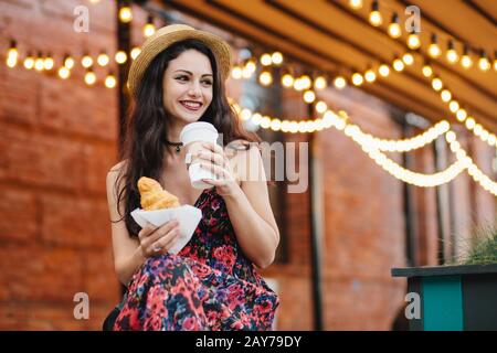 Portrait d'une femme reposante avec des cheveux sombres, des yeux brillants et des lèvres en forme de puits portant un chapeau et une robe, ayant dîner, restaurant croissant et café à boire Banque D'Images