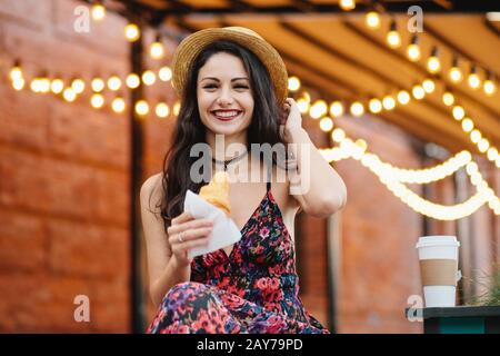 Belle femme avec des cheveux sombres, une apparence attrayante, portant un chapeau, une robe et un collier, tenant un délicieux croissant ayant un look gai. Heureux jeunes Banque D'Images