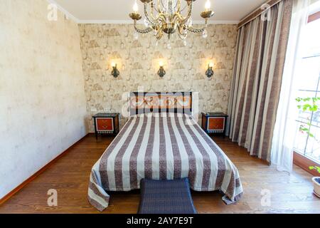 Chambre élégante et chère avec un grand lit blanc et un couvre-lit à rayures. Une tête de lit haute avec un motif rouge, des lampes murales torsadées et un lustre. B Banque D'Images