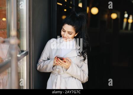 Portrait d'une femme brunette à la mode avec une apparence agréable, une belle manucure portant un imperméable blanc tenant le smartphone dans ses mains avoir le bouton Banque D'Images