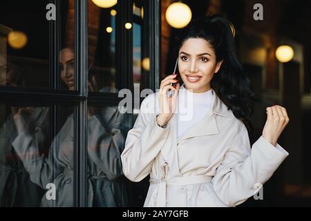 Jolie femme brunette avec des cheveux ondulés attachés dans la queue de poney ayant un bon visage pâle regardant avec élégance tout en se posant à l'extérieur à l'aide d'un gadget moderne. Mignon Banque D'Images