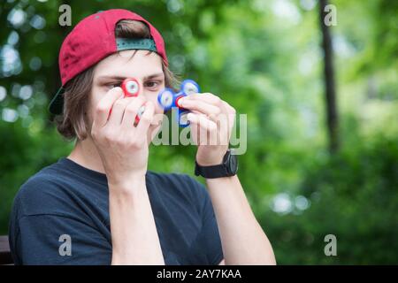 Un hipster à poil long dans une casquette est assis sur un banc et tourne un disque de fidget Banque D'Images