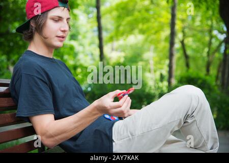 Un hipster à poil long dans une casquette est assis sur un banc et tourne un disque de fidget Banque D'Images