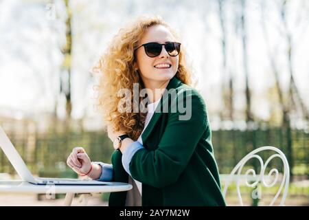 Heureuse femme élégante avec des cheveux frisés portant des lunettes de soleil travaillant avec un ordinateur portable à l'extérieur dans le parc taper les documents nécessaires en retournant en remarquant certains Banque D'Images