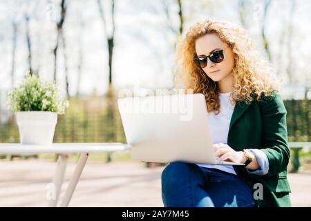 Portrait extérieur d'une jeune fille attrayante avec des cheveux blonds doux portant des lunettes de soleil, un Jean et une veste verte tenant ordinateur portable sur ses genoux lire Banque D'Images