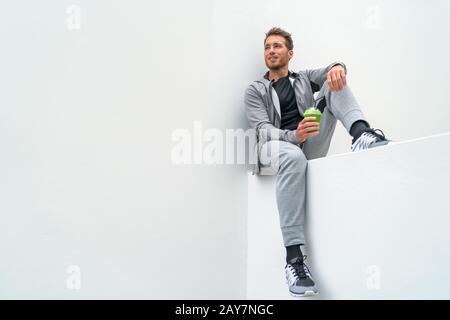 Homme de sport en bonne santé buvant un smoothie vert pendant l'entraînement à la salle de gym dans le pantalon de sueur d'activewear en plein air. Athlète assis après l'entraînement. Banque D'Images