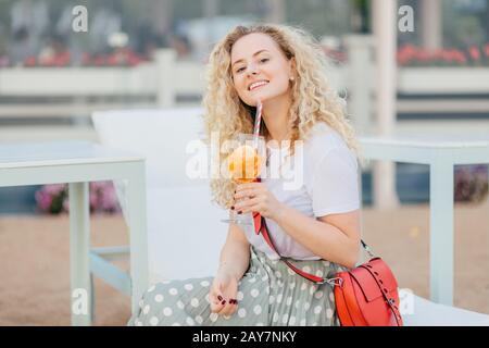 Heureuse et heureuse jeune femme curieuse vêtue d'un t-shirt blanc et d'une jupe longue, elle tient un verre de cocktail, aime le soleil jour et de bonnes vacances, a manucure, Banque D'Images