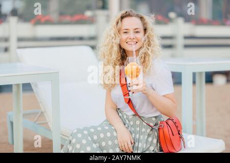 Une femme blonde heureuse aux cheveux caillés, qui tient un cocktail orange frais, est dans un esprit haut, vêtue de façon décontractée, pose en plein air sur la plage, expresse Banque D'Images