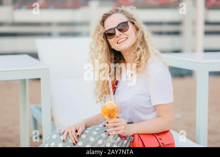 Jolie jeune femme avec des cheveux ondulés, porte des lunettes de soleil, se repose bien pendant l'été, tient des boissons fraîches, a le sourire amical, semble positivement Banque D'Images
