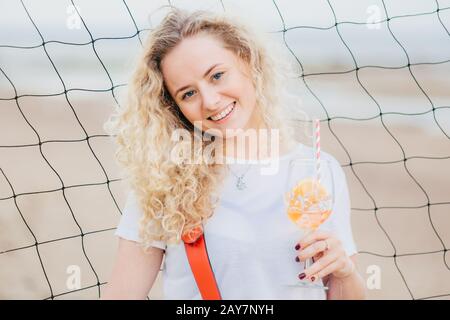 Photo horizontale de gaie heureuse blonde femme vêtue de vêtements décontractés, contient un cocktail orange frais, sourires positivement, pose contre le filet de tennis bac Banque D'Images