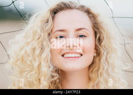 Gros plan de la jeune femme curieuse a un large sourire brillant, regarde positivement l'appareil photo, a une expression joyeuse, se tient en plein air, a une peau fraîche, Banque D'Images