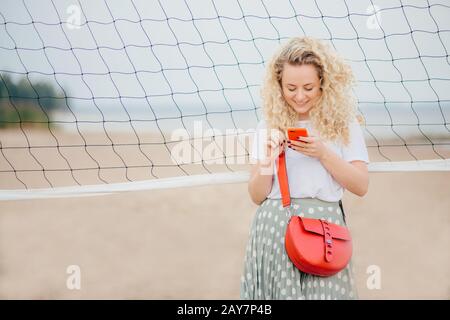 Modèle féminin attrayant concentré dans le smartphone, lit l'information sur la page Web, a un sac lumineux, sourires joyeusement comme recevoir le mesage de petit ami, pos Banque D'Images