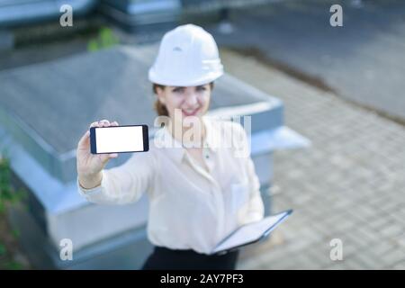 Jolie femme d'affaires en chemisier blanc, montre, casque et jupe noire se tiennent sur le toit et montrent smartphone vide Banque D'Images