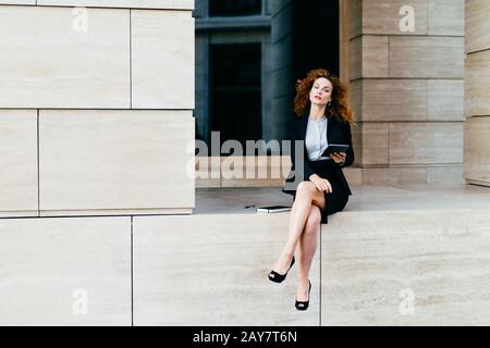 Jolie femme mausseuse avec un look assuré, assise seule, montrant ses jambes longues et minces, vêtues d'une veste noire avec jupe, tenant une tablette, lookin Banque D'Images