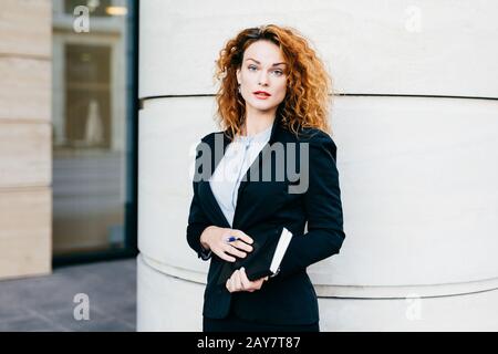 Jeune femme belle portant des vêtements formels, habillés formellement, tenant le pocketbook avec le stylo, ayant une coiffure bushy, debout près de bureau builduing. S Banque D'Images