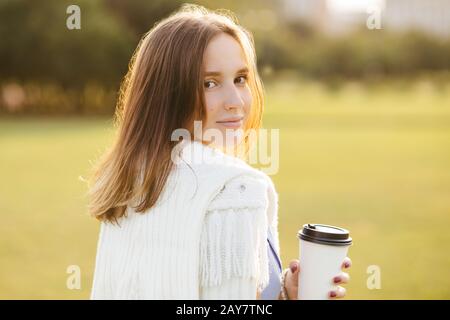 Modèle jeune femme décontracté avec cheveux sombres, boissons café à emporter, se tient à l'arrière, a une promenade en plein air, bénéficie d'une journée ensoleillée et chaude, ressemble directement à un Banque D'Images