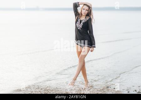 Jeune femme assez mince et en bonne santé avec de longues jambes minces se tient près de la mer calme avec de petites vagues et fond bleu, étant satisfait de poser à l'appareil photo, Banque D'Images