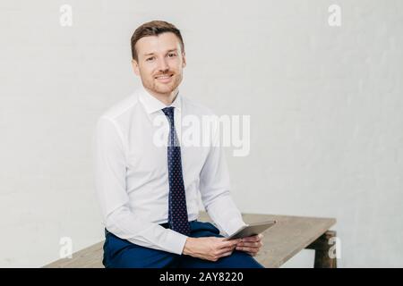 La photo intérieure du financier masculin à expression positive porte une chemise blanche classique avec cravate, tient une tablette moderne dans les mains, isolée sur blanc b Banque D'Images