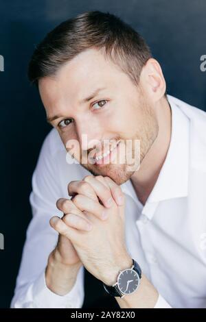 Portrait vertical d'un homme malrasé et gai avec un sourire amical, maintient les paumes enfoncées sous le menton, porte la montre à portée de main, semble heureux Banque D'Images