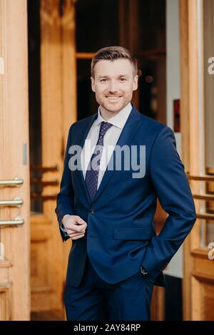 La photo verticale du bel employé masculin non rasé a une expression gaie, vêtue d'un costume élégant, se tient près de l'entrée de son entreprise, étant en bon état Banque D'Images