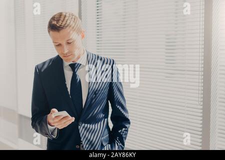 Jeune entrepreneur réussi en costume cher, tient le téléphone mobile, se tient au bureau, contre la fenêtre dans le cabinet, chats en ligne, lit des nouvelles sur finan Banque D'Images