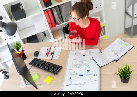 Une jeune fille est assise au bureau, tenant une tasse rouge dans ses mains et regardant le moniteur. Une carte magnétique l Banque D'Images