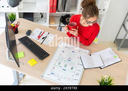 Une jeune fille est assise à la table dans le bureau, tenant une tasse rouge dans ses mains et regardant le bloc-notes. Une carte magnétique Banque D'Images