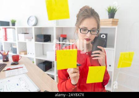 Une jeune fille se tient près d'une administration transparente avec autocollants et est titulaire d'un verre au café et un marqueur. Banque D'Images