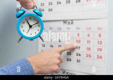 Une main tient un petit réveil, dans l'arrière-plan d'une part de points de longs week-ends et jours fériés sur un calendrier mural Banque D'Images