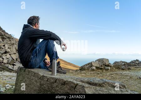 Homme assis sur la roche dans les montagnes, boire du café et regarder vers le bas sur la mer en dessous Banque D'Images