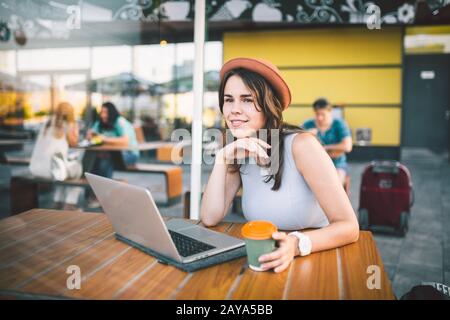 Belle jeune femme caucasienne assis sur une terrasse dans un café en été à une table en bois dans un chapeau et une plaque utilise la technologie, Banque D'Images