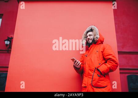 beau jeune homme étudiant avec un sourire toothy et des supports de barbe sur fond de mur rouge dans une veste d'hiver rouge vif avec capuche avec Banque D'Images