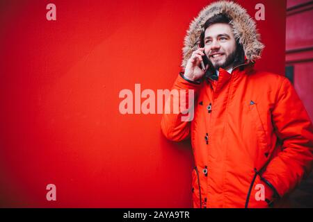 beau jeune homme étudiant avec un sourire toothy et une barbe se tient sur un fond de mur rouge dans une veste d'hiver rouge vif avec un Banque D'Images