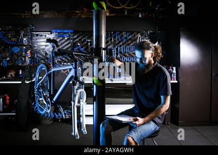 Jeune homme barbu élégant avec de longs cheveux homme caucasien homme d'affaires homme d'affaires travailleur dans l'atelier de vélo utilise le portable pour wr Banque D'Images
