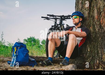 vélo de montagne masculin reposant sur une promenade en vélo, assis sur le sol sous un arbre avec son vélo de montagne, se tient à côté de lui, appréciant th Banque D'Images