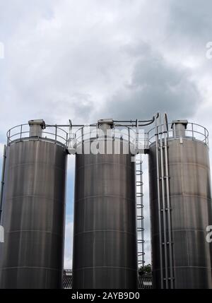 un groupe de trois grands réservoirs de stockage de produits chimiques en acier avec tuyaux de raccordement et échelles contre un ciel bleu nuageux Banque D'Images