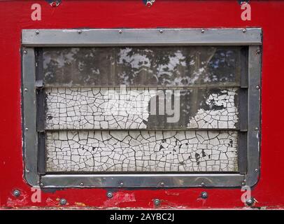 une vieille fenêtre de caravane affligée avec cadre en acier et fenêtres claquées avec une vieille peinture blanche fissurée avec un cadre rouge avec rivets Banque D'Images